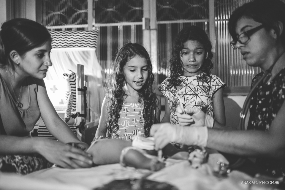 Irmãs se preparam para cortar o cordão umbilicam do bebê recém nascido. Uma delas faz cara de assustada para o bisturi afiado.