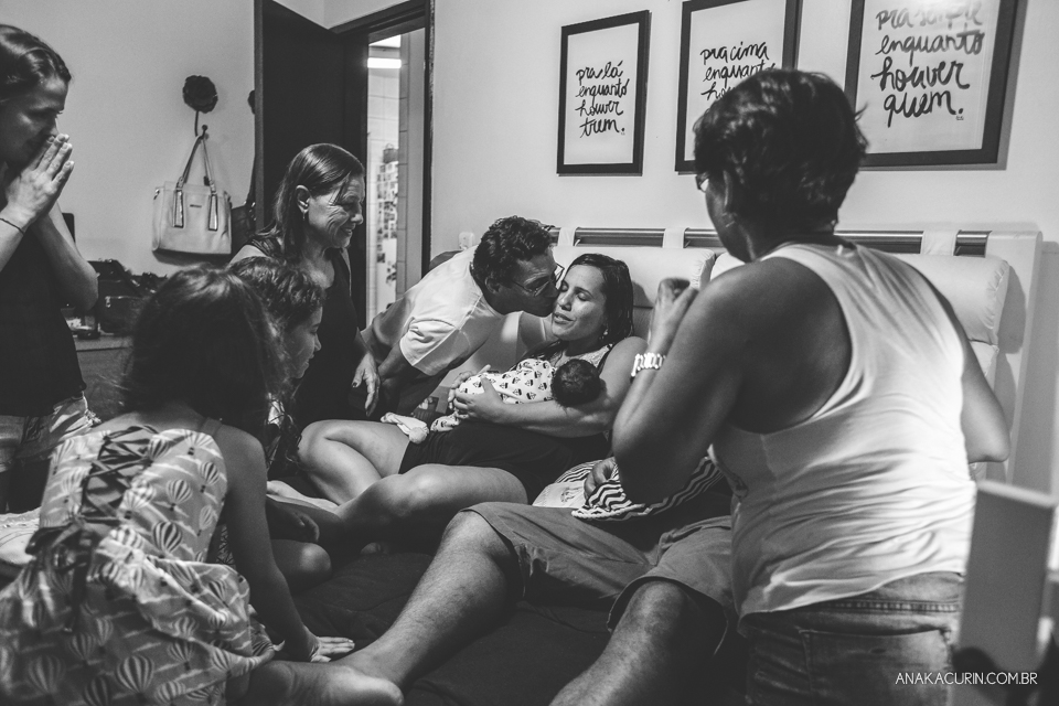 Família reunida em volta de mulher recém parida com filho no colo na cama do casal. Pai beija a filha.