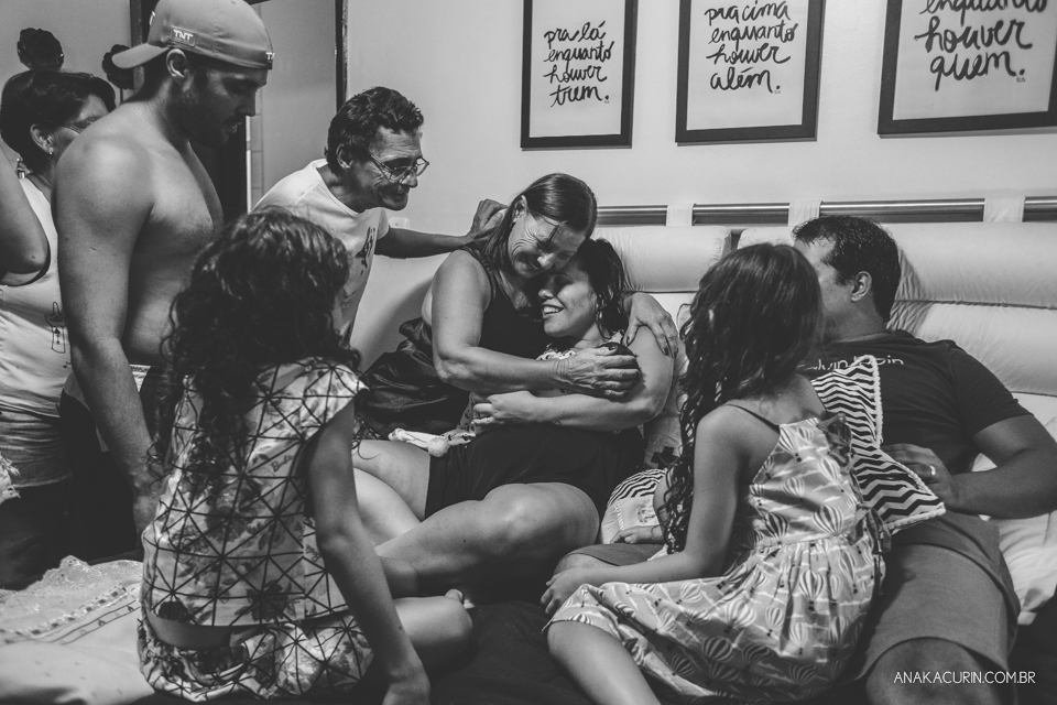 Família reunida em volta de mulher recém parida com filho no colo na cama do casal. Mãe da mulher a abraça.