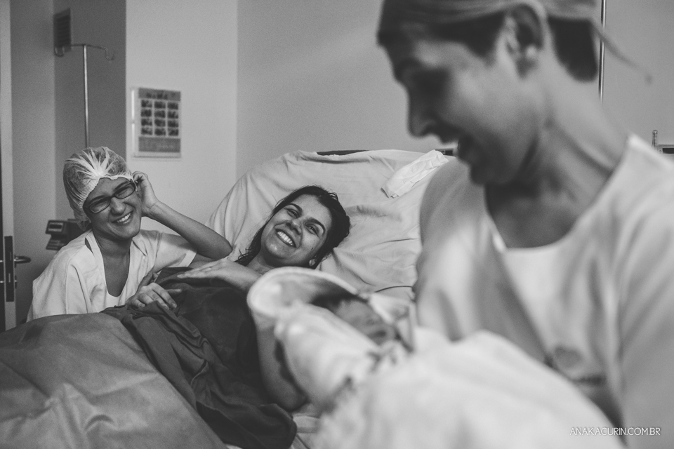 Parto natural hospitalar de Sofia, que aconteceu na Perinatal de Laranjeiras e teve a fotografia feita por Ana Kacurin. Doula e mãe sorriem enquanto olham pai segurando bebê.