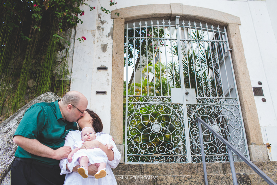 Batizado da Gabi, que aconteceu na igraja Nossa Senhora do Outeiro da Glória, no Rio de Janeiro. Pais se beijam com bebê no colo em frente a um portão de ferro bonito.