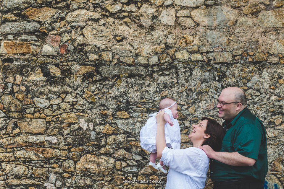 Batizado da Gabi, que aconteceu na igraja Nossa Senhora do Outeiro da Glória, no Rio de Janeiro. Pais levantam a bebê na frente de uma parede de pedra.