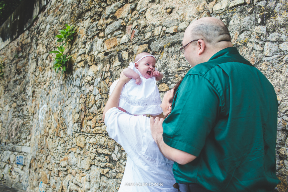 Batizado da Gabi, que aconteceu na igraja Nossa Senhora do Outeiro da Glória, no Rio de Janeiro. Pais levantam a bebê na frente de uma parede de pedra.
