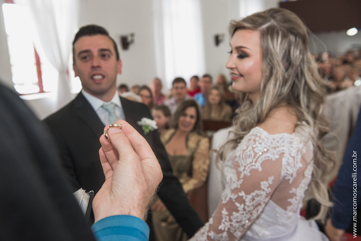 Fotografia das alinças de casamento na mão do pastor com os noivos desfocados ao fundo. Foto por Marco Moscarelli
