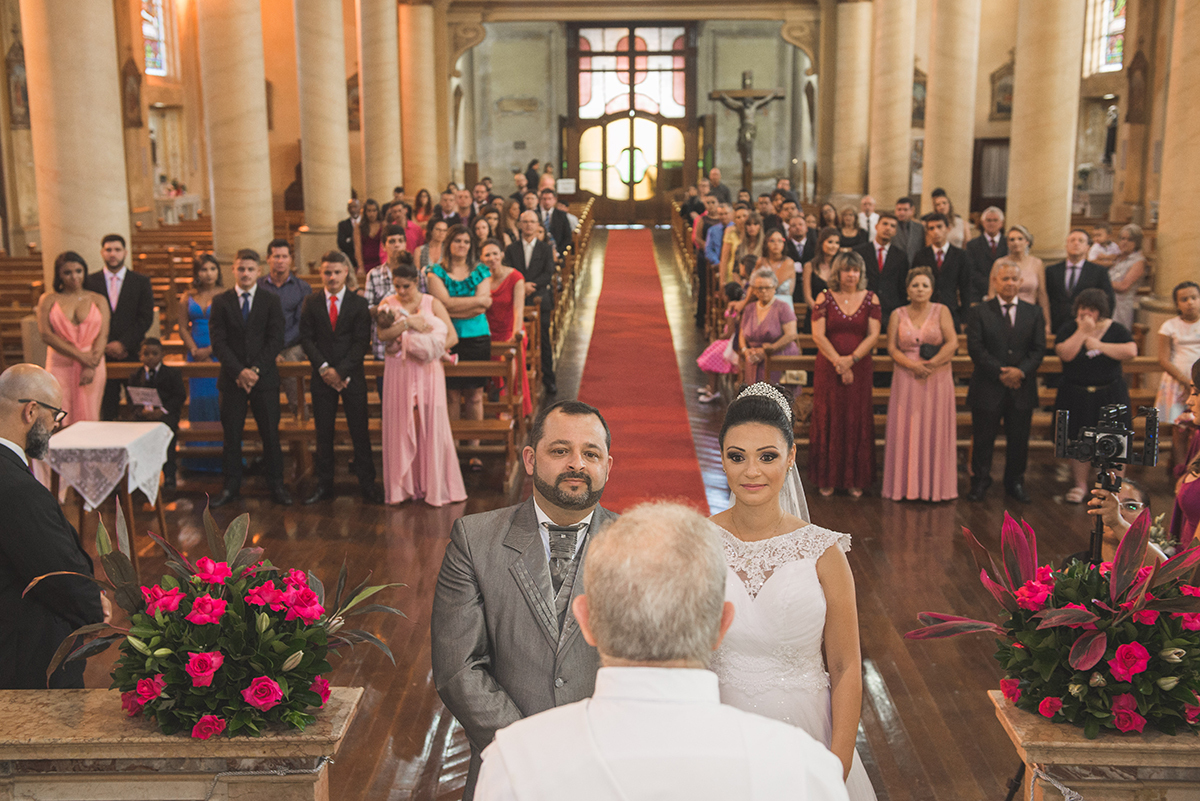 Fotografia dos noivos no altar durante a cerimônia de casamento com a igreja e convidados ao fundo. Foto por Marco Moscarelli