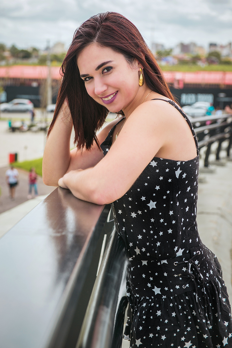 Linda foto da aniversariante no Parque UNa com um vestido preto debruçada em corrimão de passarela sorrindo. Foto por Marco Moscarelli