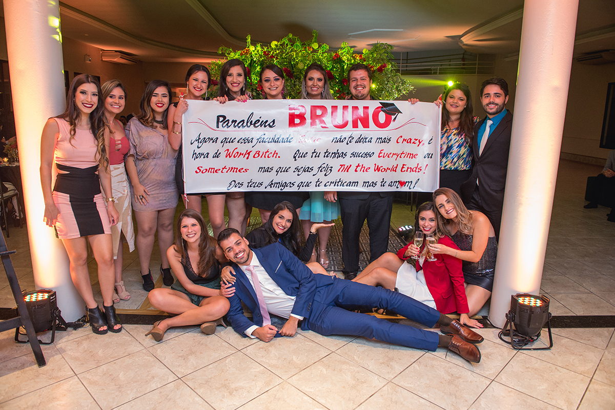 Formando deitado no chão a frente de uma faixa de homenagem com suas convidadas durante a sua recepção de formatura em direito em Pelotas. Foto por Marco Moscarelli Fotografo.