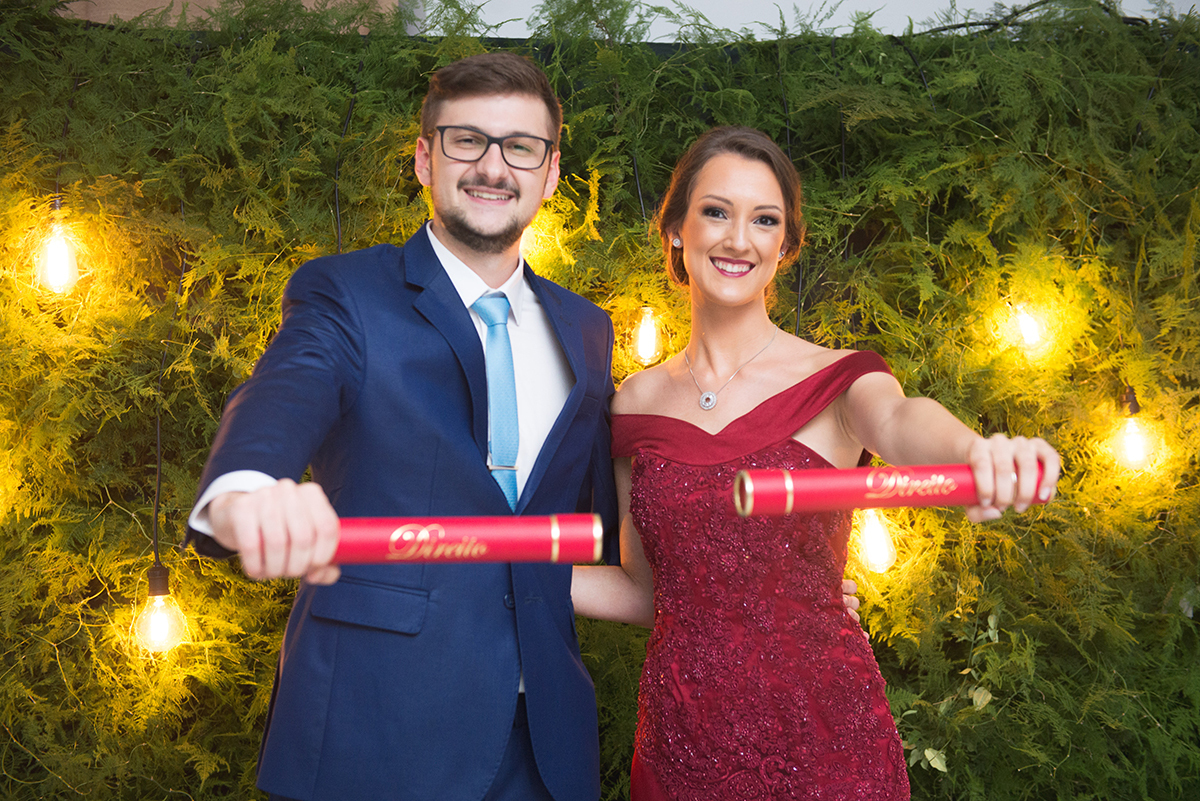 Casal de noivos e formandos posando para fotos no painel com seus respectivos canudos de formatura. Foto por Marco Moscarelli Fotografo.