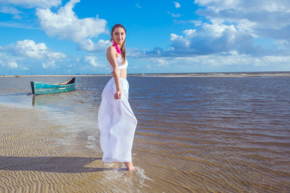 Linda foto da debutante caminhando pela praia com um pequeno barco ao fundo  durante seu ensaio de 15 anos na praia da capilha. Foto por Marco Moscarelli Fotografo.