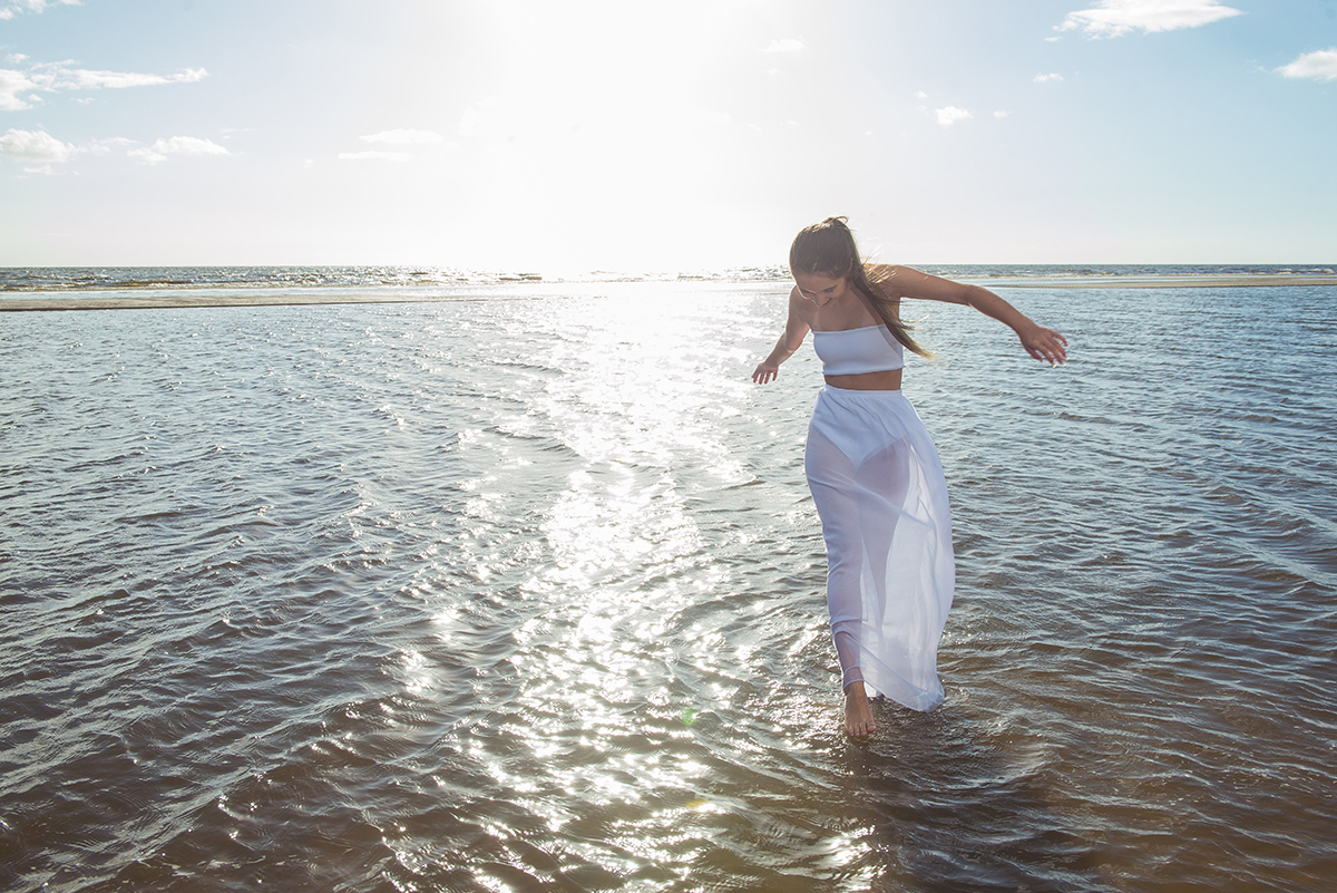 Linda foto da debutante caminhando pela praia com um pequeno barco ao fundo  durante seu ensaio de 15 anos na praia da capilha. Foto por Marco Moscarelli Fotografo.