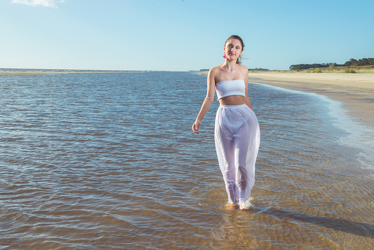 Linda foto da debutante caminhando pela praia com um pequeno barco ao fundo  durante seu ensaio de 15 anos na praia da capilha. Foto por Marco Moscarelli Fotografo.