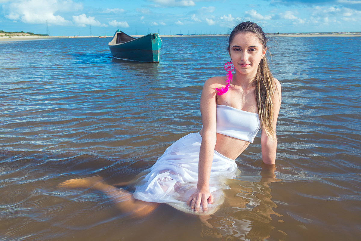 Linda foto da debutante sentada na água com um sorriso lindo e  com um pequeno barco ao fundo  durante seu ensaio de 15 anos na 