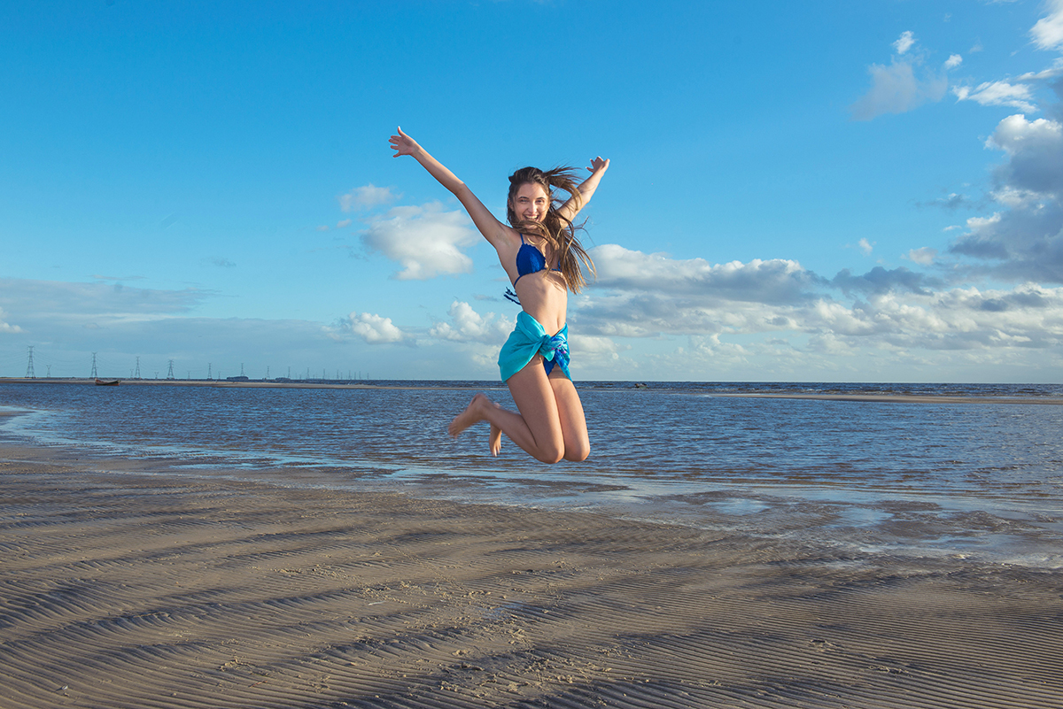 Fotografia da debutante com um lindo biquíni azul saltando alegremente na praia da capilha. Foto por Marco Moscarelli Fotografo 