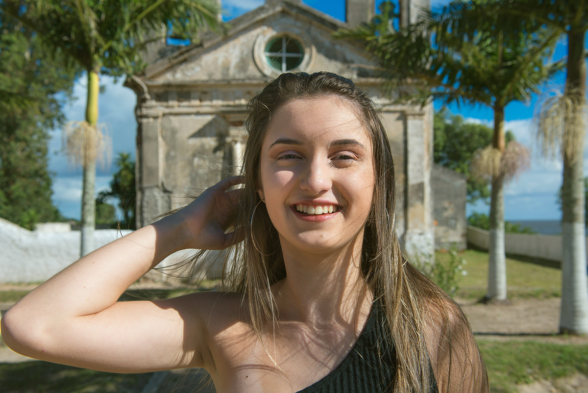Fotografia da debutante com a mão no cabelo com uma pequena igreja ao fundo durante o seu ensaio fotográfico na praia da capilha. Foto por Marco Moscarelli Fotografo.