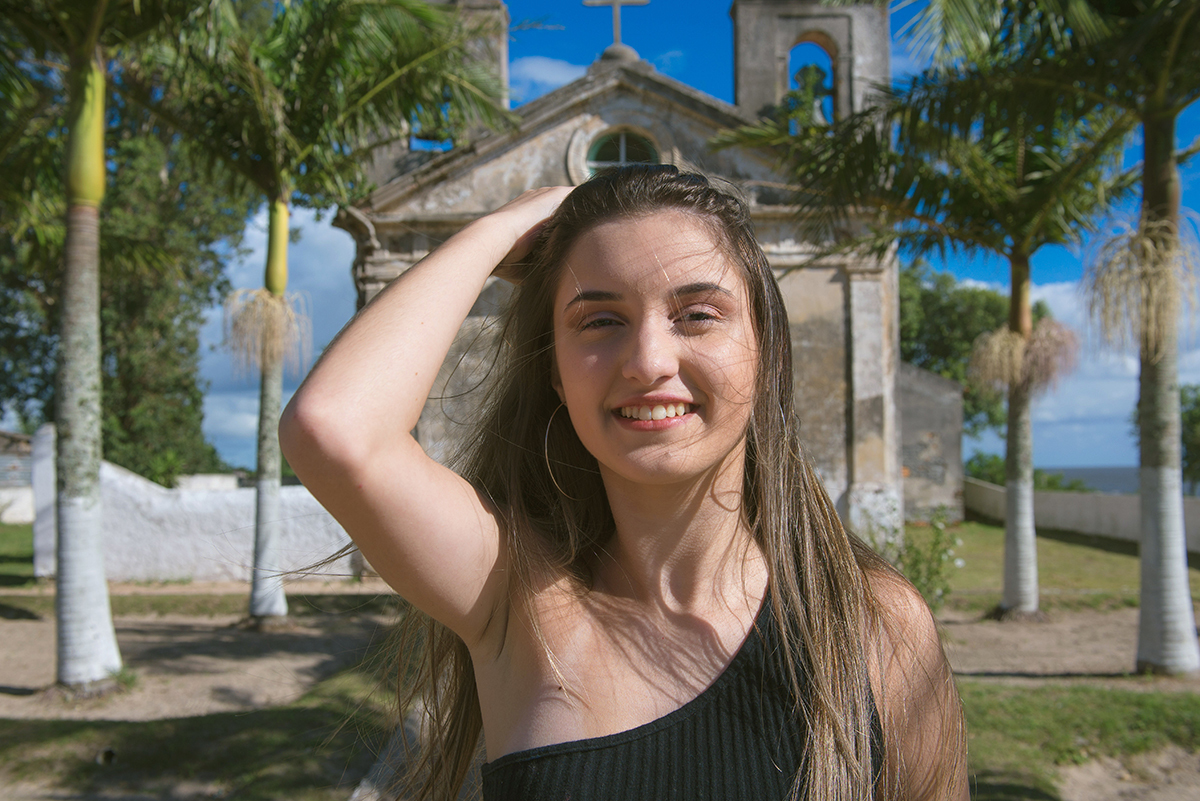 Fotografia da debutante com a mão no cabelo com uma pequena igreja ao fundo durante o seu ensaio fotográfico na praia da capilha. Foto por Marco Moscarelli Fotografo.
