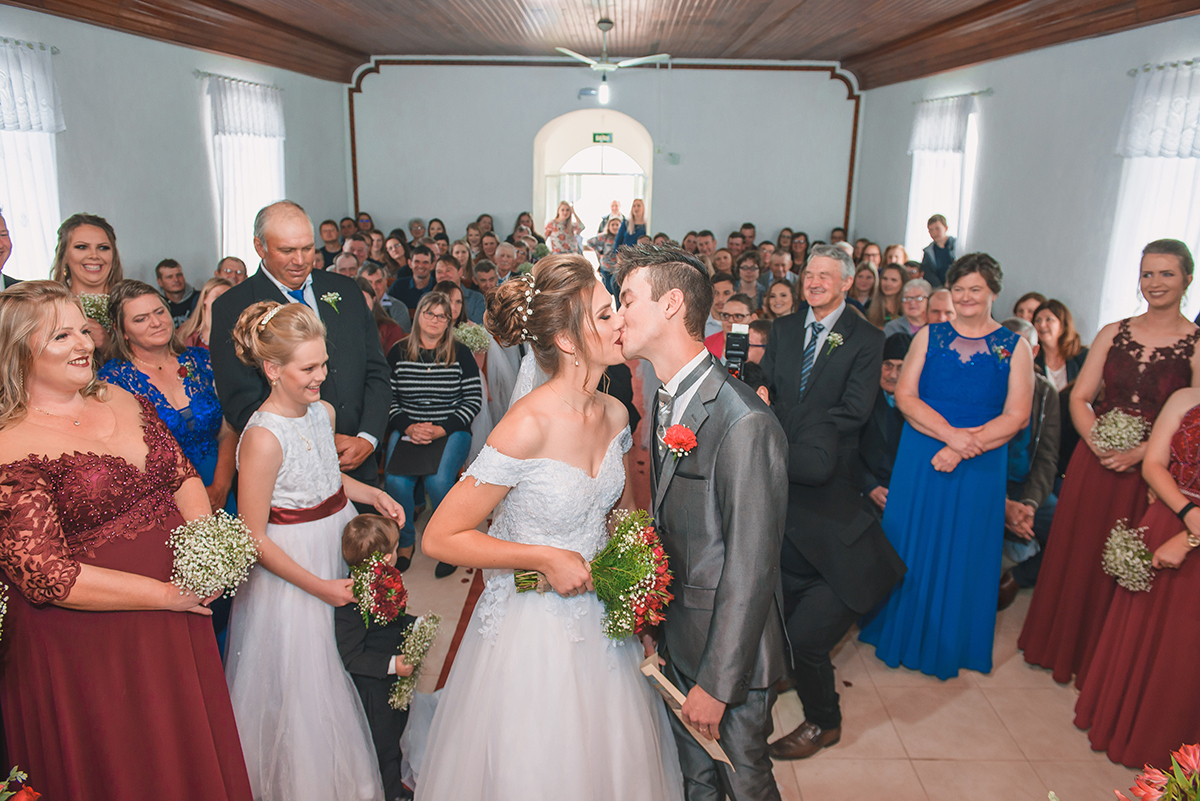 Foto do beijo dos noivos no final do casamento com padrinhos e convidados emocionados.  Foto por Marco Moscarelli Fotografo Pelotas.