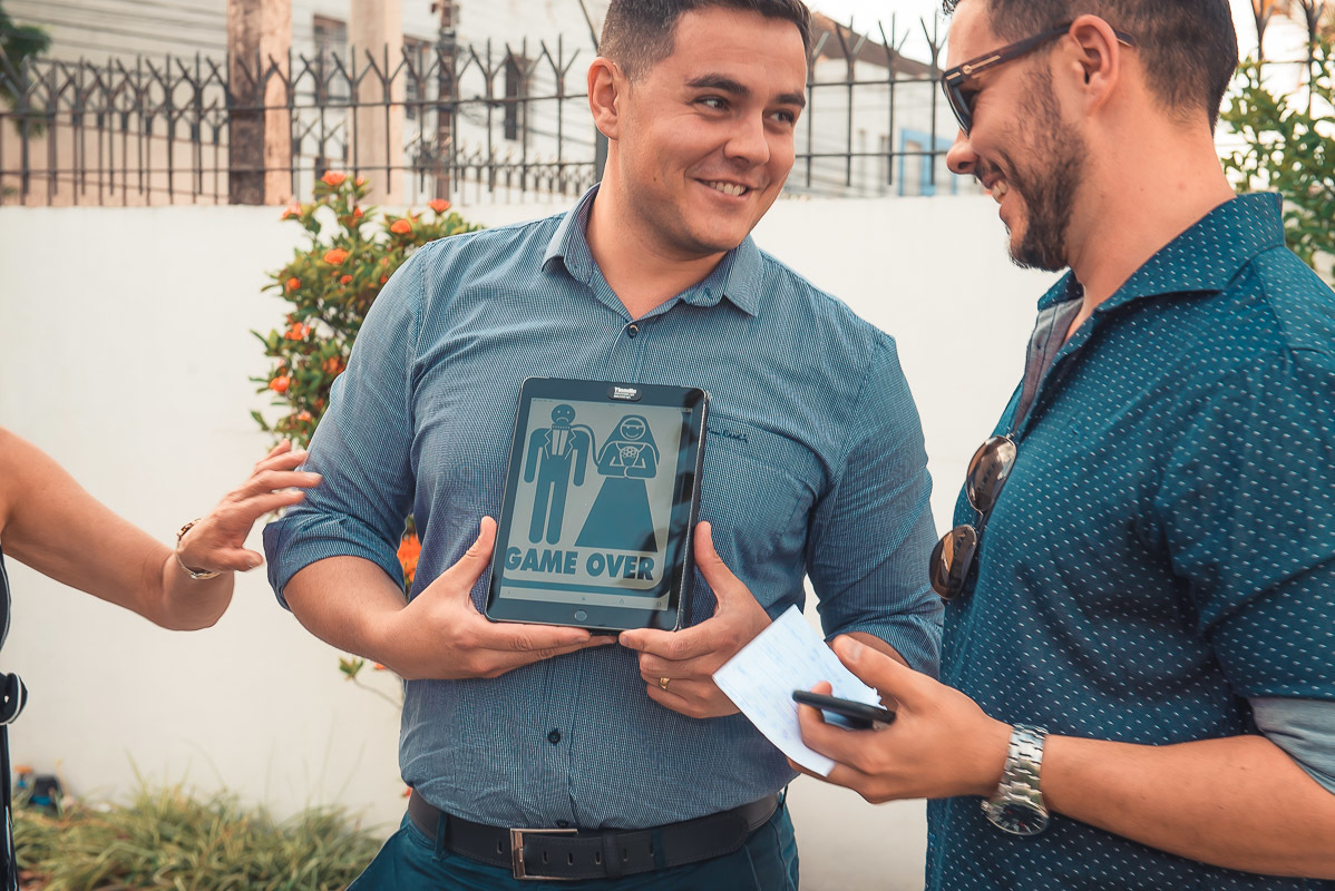 Convidados rindo alegremente com placa  brincando com noivo. Retoques finais da maquiagem da noiva momentos antes do casamento. Foto por Marco Moscarelli Fotógrafo Pelotas.