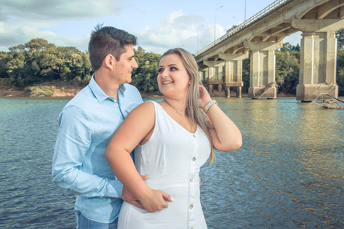 Casal de noivos posando para foto com ponte e rio ao fundo durante o seu ensaio fotográfico pré wedding. ensaio pré-wedding. ensaio de casal. Noivos trocando um belo beijo durante o seu ensaio de casal. Foto por Marco Moscarelli Fotógrafo Pelotas.
