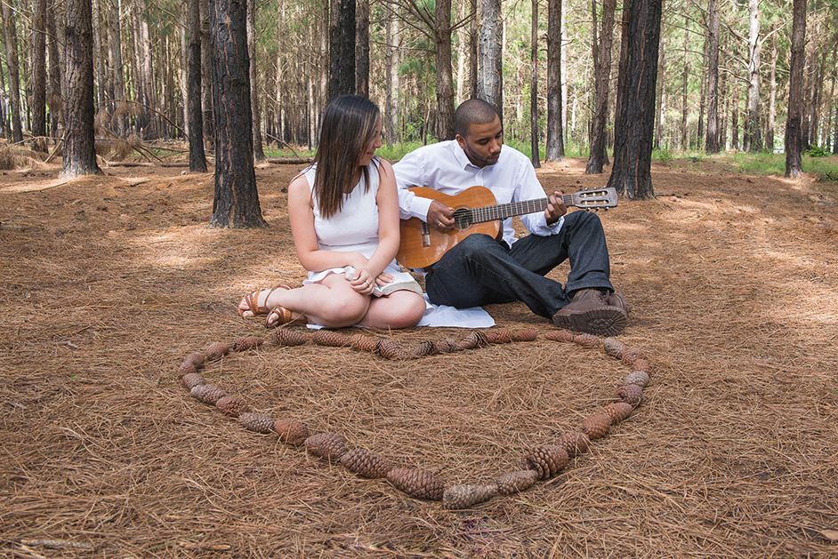 Foto do ensaio do casal, noivo tocando violão para a noiva em um bosque com um coração de pinhas ao chão.