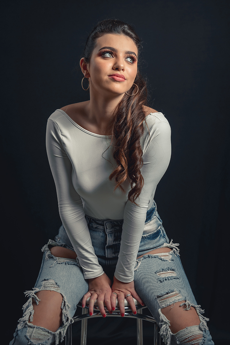 Modelo posando em estúdio com fundo preto usando uma blusa branca e calça jeans, sentada em um banco. Olhos verdes chamando atenção neste lindo editorial de moda em estúdio fotográfico. Foto por Marco Moscarelli Fotógrafo Pelotas.