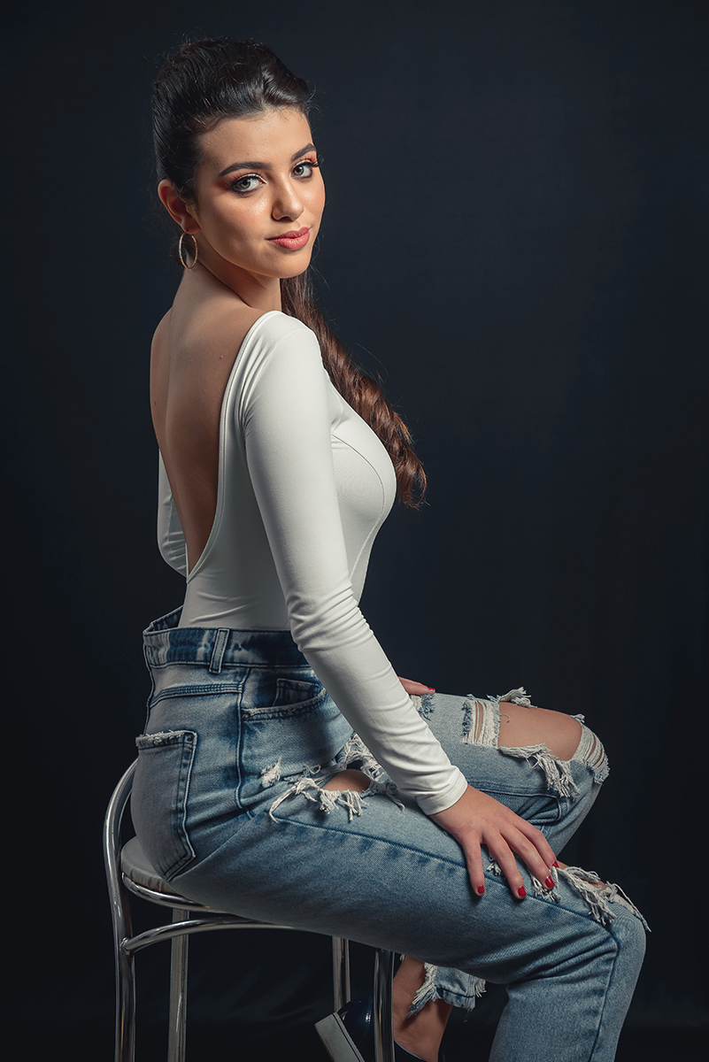 Modelo posando em estúdio com fundo preto usando uma blusa branca e calça jeans, sentada em um banco. Olhos verdes chamando atenção neste lindo editorial de moda em estúdio fotográfico. Foto por Marco Moscarelli Fotógrafo Pelotas.