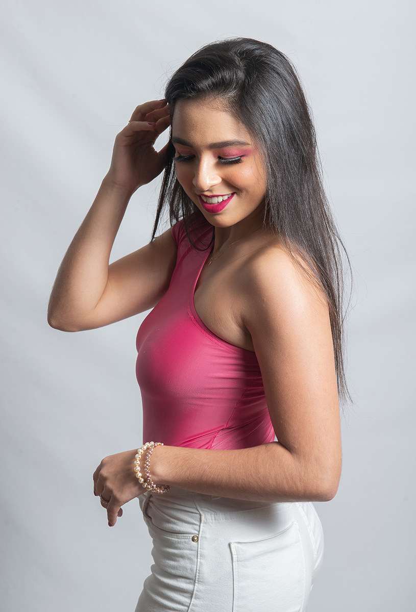 Modelo posando em estúdio com fundo branco usando uma blusa rosa e calça jeans branca. Olhos verdes chamando atenção neste lindo editorial de moda  