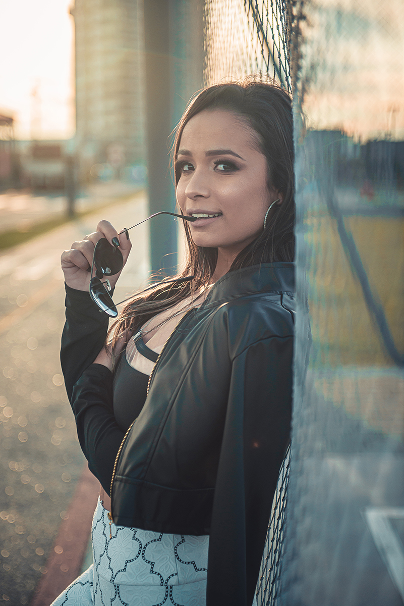 Modelo posando  encostada em tela de quadra de esportes, usando roupas despojadas e com óculos na boca  em ensaio fotográfico realizado no parque Una em Pelotas. Foto por Marco Moscarelli Fotógrafo Pelotas.