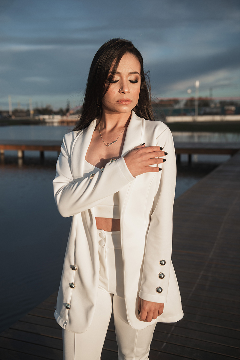 Modelo posando com a mão no cabelo e usando um terninho branco para fotografia com o fundo desfocado em ensaio fotográfico realizado no parque Una em Pelotas. Foto por Marco Moscarelli Fotógrafo Pelotas.