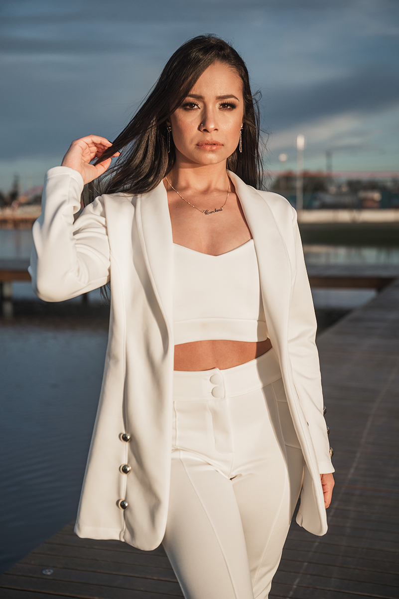 Modelo posando com a mão no cabelo e usando um terninho branco para fotografia com o fundo desfocado em ensaio fotográfico realizado no parque Una em Pelotas. Foto por Marco Moscarelli Fotógrafo Pelotas.