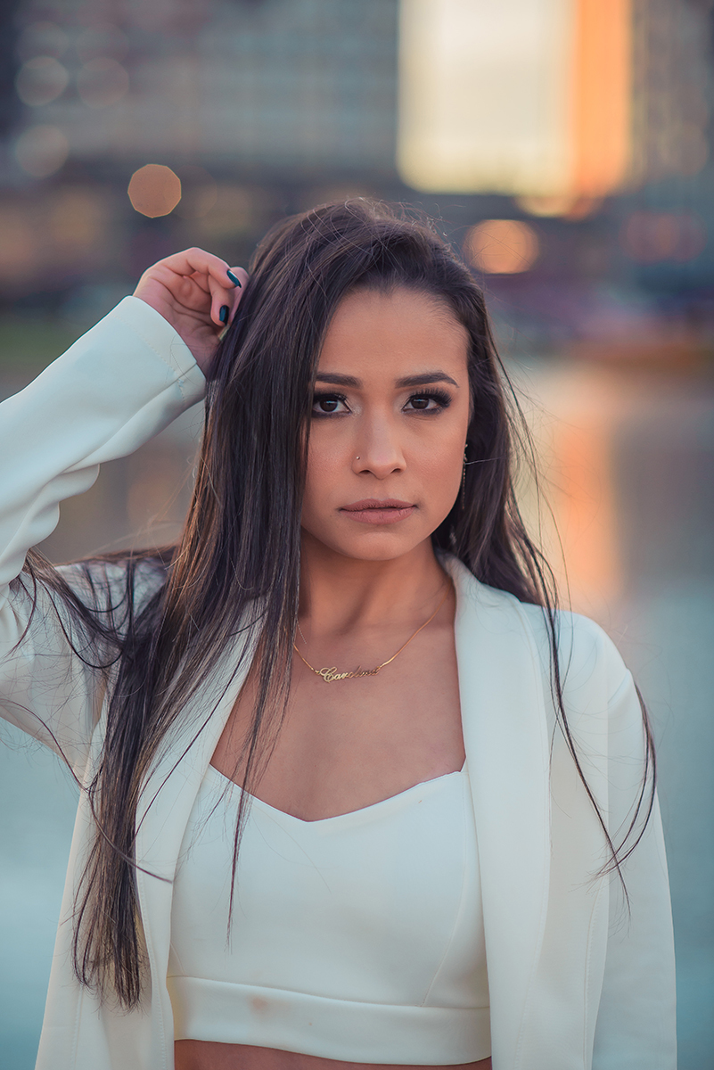 Modelo posando com a mão no cabelo e usando um terninho branco para fotografia com o fundo desfocado em ensaio fotográfico realizado no parque Una em Pelotas. Foto por Marco Moscarelli Fotógrafo Pelotas.