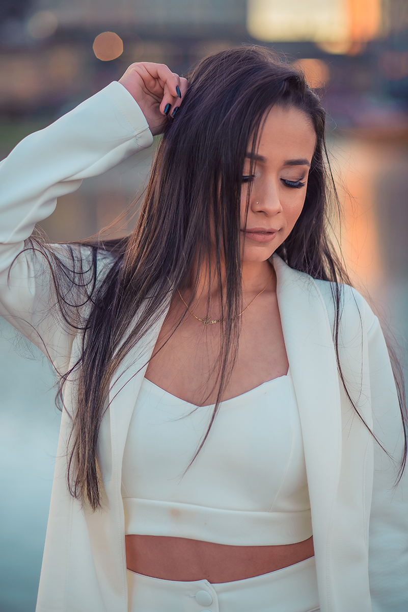 Modelo posando com a mão no cabelo e usando um terninho branco para fotografia com o fundo desfocado em ensaio fotográfico realizado no parque Una em Pelotas. Foto por Marco Moscarelli Fotógrafo Pelotas.
