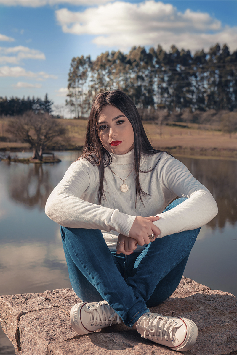 Linda foto da aniversariante sentada sobre uma pedra com o lago ao fundo, durante seu ensaio de 15 anos para seu aniversário de 15 anos. Foto por Marco Moscarelli Fotógrafo Pelotas.  
