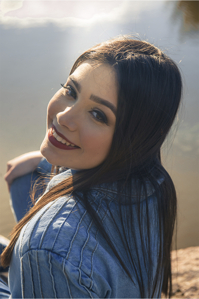Debutante sentada em uma ponde de pedras com o lago as suas costas sorrindo para a câmera, durante seu ensaio de 15 anos para seu aniversário de 15 anos. Foto por Marco Moscarelli Fotógrafo Pelotas.  
