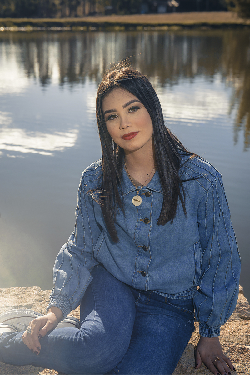 Debutante sentada em uma ponde de pedras com o lago as suas costas sorrindo para a câmera, durante seu ensaio de 15 anos para seu aniversário de 15 anos. Foto por Marco Moscarelli Fotógrafo Pelotas.  