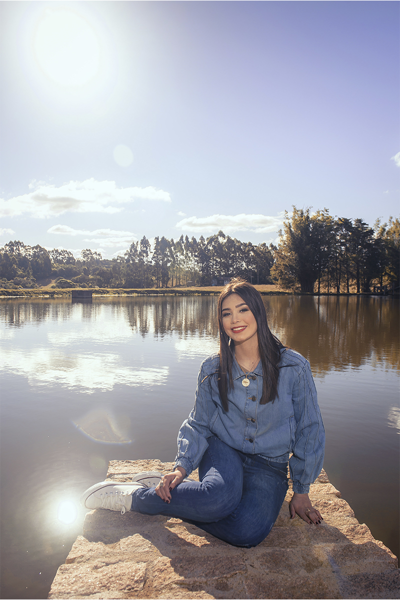 Debutante sentada em uma ponde de pedras com o lago as suas costas sorrindo para a câmera, durante seu ensaio de 15 anos para seu aniversário de 15 anos. Foto por Marco Moscarelli Fotógrafo Pelotas.  