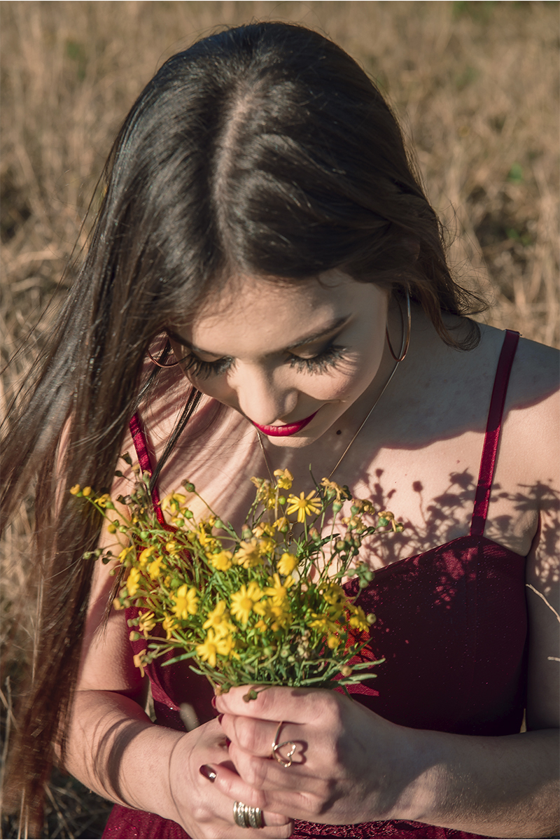 Debutante posando para foto com um buquê de flores amarelo em um campo amarelo com um lindo vestido vermelho e olhando para o sol, durante seu ensaio de 15 anos para seu aniversário de 15 anos. Foto por Marco Moscarelli 