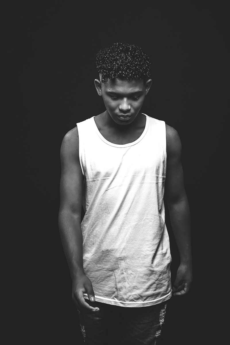 Modelo posando para foto preto e branco  em estúdio com fundo preto, usando camiseta banca e bermuda. Foto por Marco Moscarelli Fotografo Pelotas.