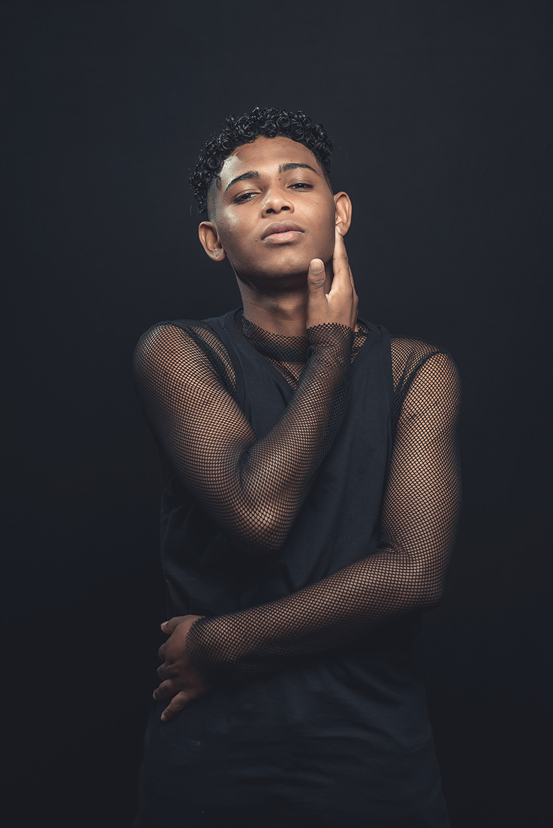 Modelo posando para foto colorida em estúdio com fundo preto, usando camiseta camisa e calça preta. Foto por Marco Moscarelli Fotografo Pelotas