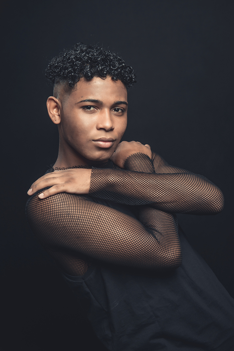 Modelo posando para foto colorida em estúdio com fundo preto, usando camiseta camisa e calça preta. Foto por Marco Moscarelli Fotografo Pelotas