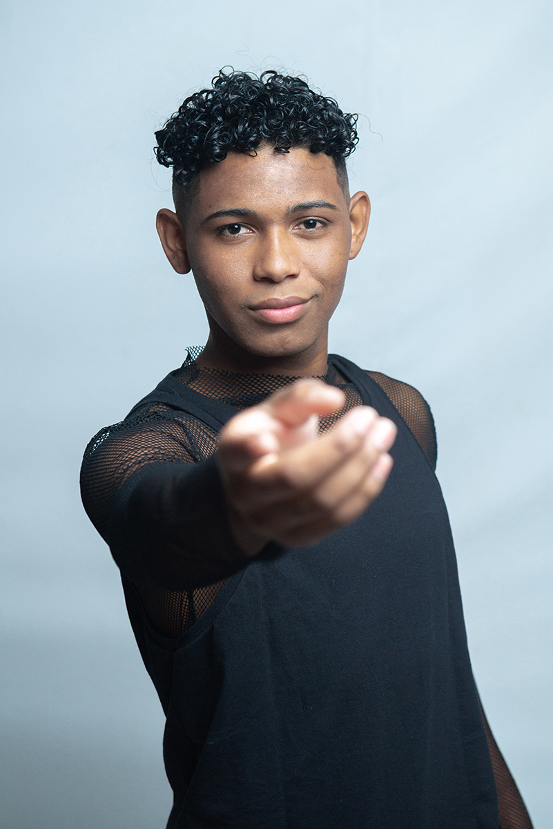 Modelo posando para foto colorida em estúdio com fundo branco, usando camiseta camisa e calça preta. Foto por Marco Moscarelli Fotografo Pelotas