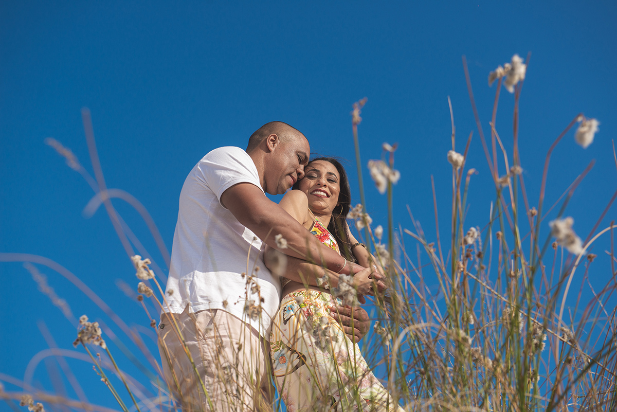 Fotografia do casal em seu pré wedding na praia com o céu azul ao fundo e flores compondo a foto no ensaio de casal. Foto por Marco Moscarelli Fotógrafo Pelotas.