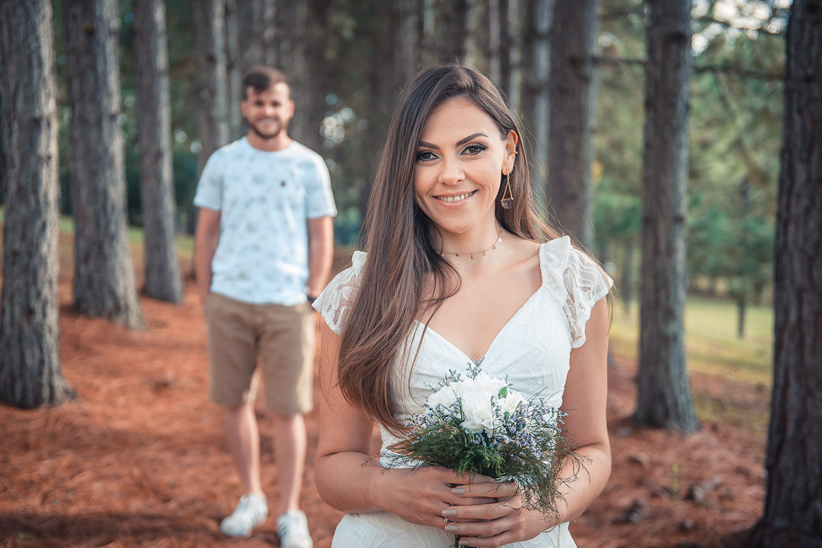 Fotografia da noiva em primeiro plano com o noivo desfocado atrás, durante seu ensaio pré wedding no seu pré casamento na chácara Fiss. Foto por Marco Moscarelli Fotógrafo Pelotas.