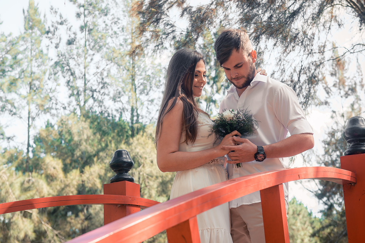 Noivo posando com a noiva em uma ponte em meio a natureza, admirando o buquê da noiva   durante seu ensaio pré wedding no seu pré casamento na chácara Fiss. Foto por Marco Moscarelli Fotógrafo Pelotas.