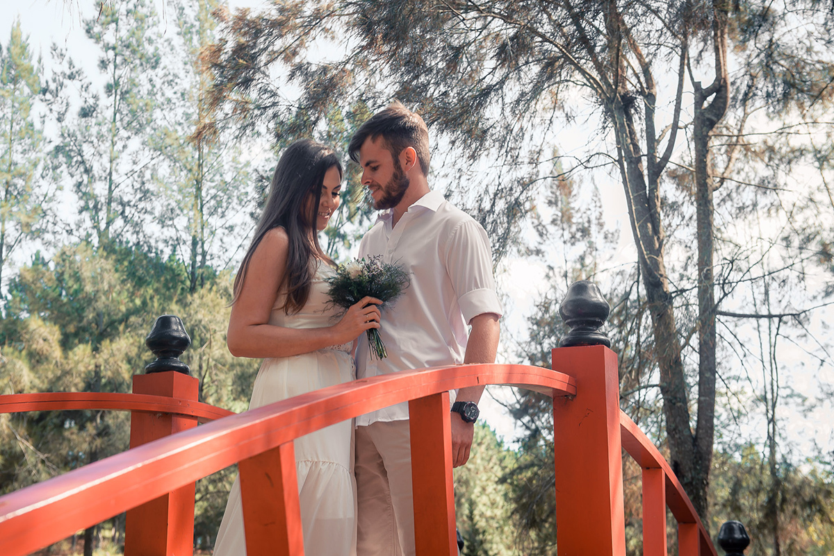 Noivo posando com a noiva em uma ponte em meio a natureza, admirando o buquê da noiva   durante seu ensaio pré wedding no seu pré casamento na chácara Fiss. Foto por Marco Moscarelli Fotógrafo Pelotas.