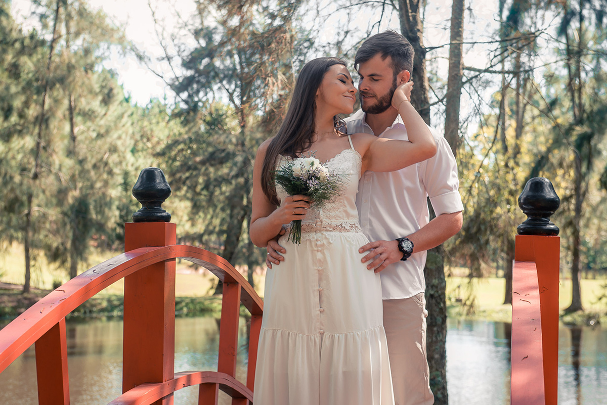 Noivo posando com a noiva em uma ponte em meio a natureza, admirando o buquê da noiva   durante seu ensaio pré wedding no seu pré casamento na chácara Fiss. Foto por Marco Moscarelli Fotógrafo Pelotas.