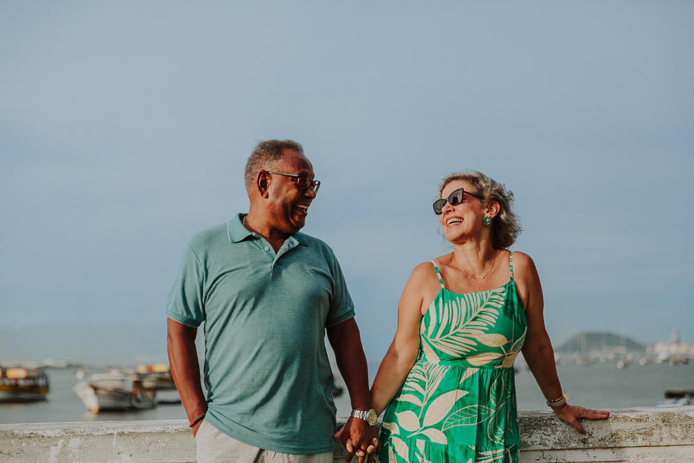 Fotografia afetiva de casal maduro rindo juntos com o mar e barcos ao fundo no Rio de Janeiro.