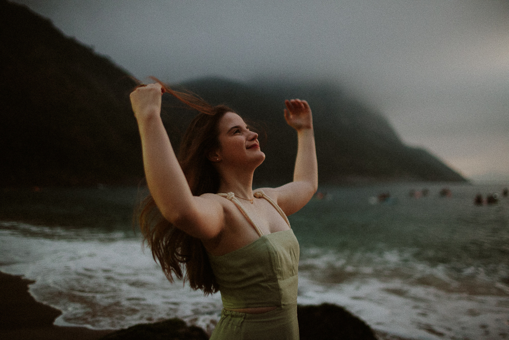 Mulher sorrindo com o vento nos cabelos durante ensaio fotográfico na praia em dia nublado em um retrato poetico