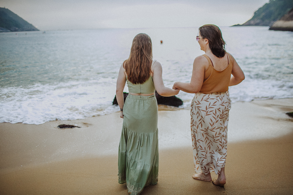 Mãe e filha caminhando pela praia ao nascer do sol em dia nublado.