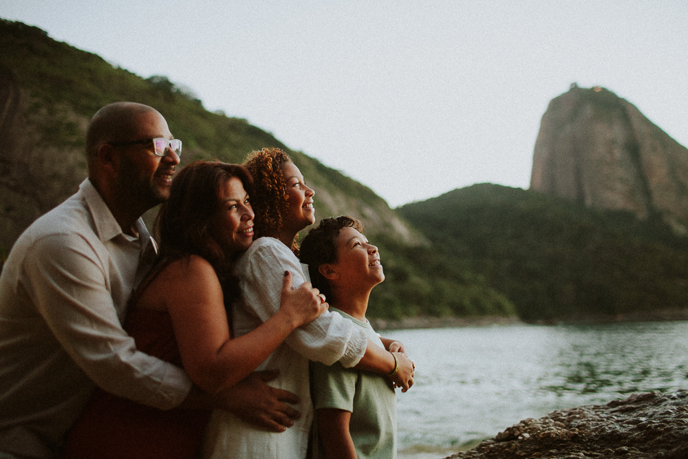 Família reunida e abraçada diante do Pão de Açúcar durante ensaio de 15 anos na Praia Vermelha, Rio de Janeiro.
