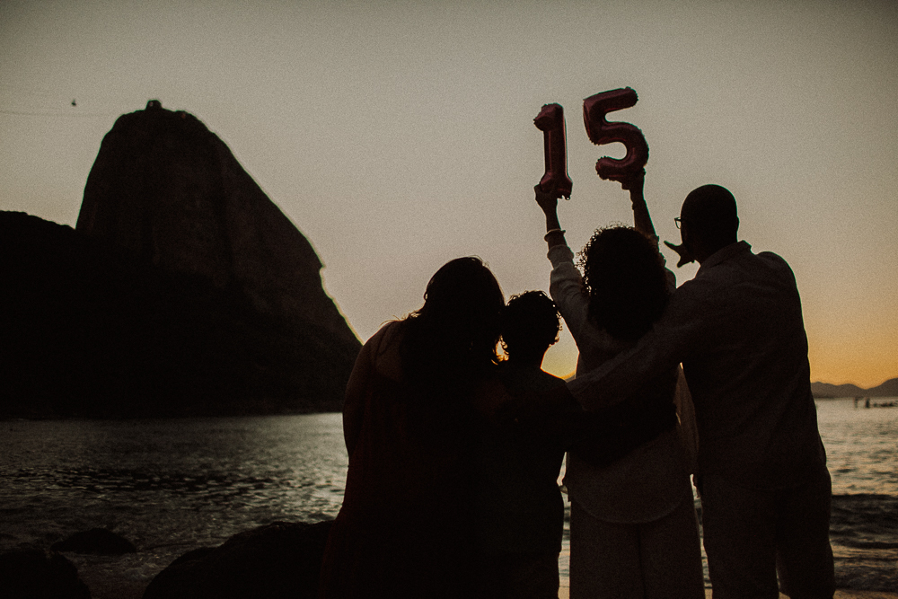 Família em silhueta celebrando os 15 anos de Alice ao pôr do sol na Praia Vermelha, com o Pão de Açúcar ao fundo.
